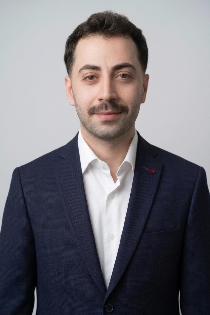 Porträt eines Mannes mit Schnurrbart in dunkelblauem Sakko und weißem Hemd vor hellem Hintergrund.